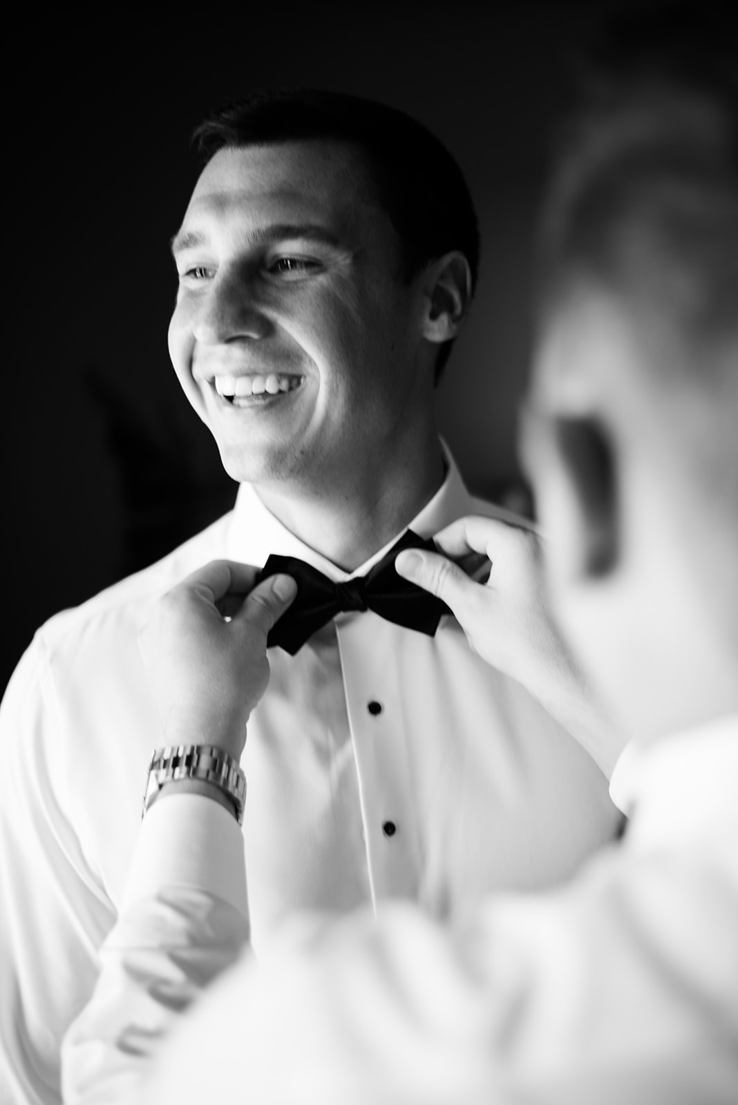 Black and white film portrait of a smiling groom having his black bow tie adjusted by a friend.